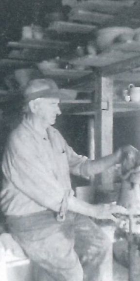 Mężczyzna pracuje przy kole garncarskim. Wokół na półkach gliniane naczynia.