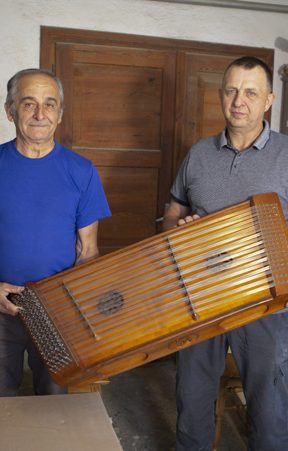 Dwóch mężczyzn trzyma w rękach duży, prostokątny, drewniany instrument.