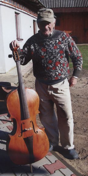 Mężczyzna w czapce z daszkiem, swetrze i jasnych spodniach stoi na podwórku. W prawej ręce trzyma duży instrument muzyczny-basy.