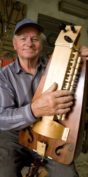 Mężczyzna w koszuli w pasy trzyma w rękach duży, drewniany instrument - lirę korbową.